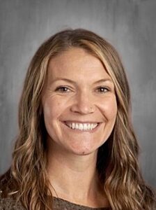 Smiling woman with wavy brown hair, wearing a brown sweater, against a gray background.