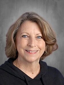 Smiling woman with shoulder-length brown hair, wearing a black hoodie, against a gray background.