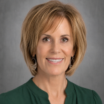 A woman with short, light brown hair smiles warmly, wearing a green blouse and silver earrings against a gray background.