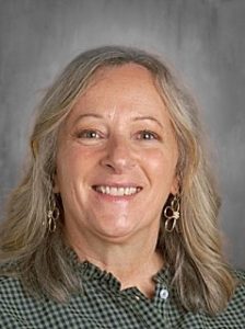 Smiling woman with shoulder-length hair, wearing hoop earrings and a checkered top, in front of a gray background.