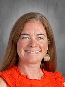 A smiling woman with long brown hair, wearing an orange top and earrings, against a gray background.