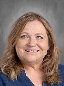 Smiling woman with shoulder-length wavy hair, wearing a blue shirt, against a gray background.