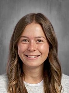 A smiling young woman with long, wavy hair, wearing a light gray sweater, against a neutral background.