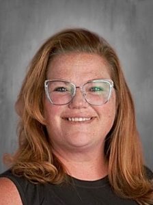 A person with shoulder-length hair and glasses smiles against a gray background.
