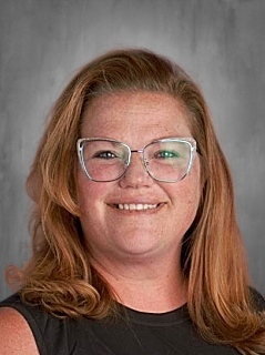 A person with shoulder-length hair and glasses smiles against a gray background.