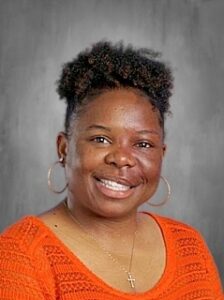 Smiling woman with short curly hair, wearing an orange shirt and hoop earrings, against a gray background.