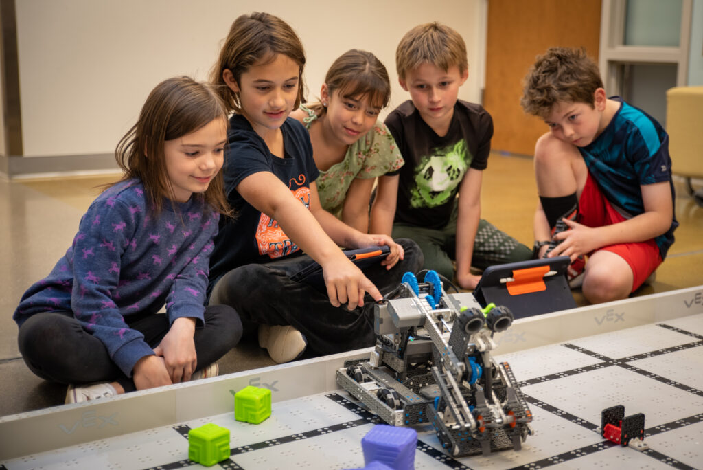 Five 4th and 5th grade girls sitting on the ground in a row, three girls on the left and two boys on the right. The second girl in from the left is pointing at the robot they made which is on a grid on the ground in front of all of the students. The rest of the students are looking at it while she points. 