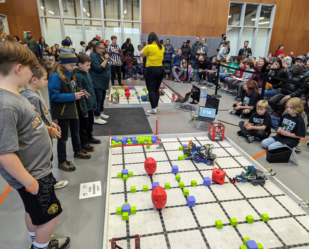 Students from Mead and another school standing on the left of a grid which is on the ground where robots that students built are competing. Students from another school are sitting on the ground on the right. A crowd is seated in chairs around them. 