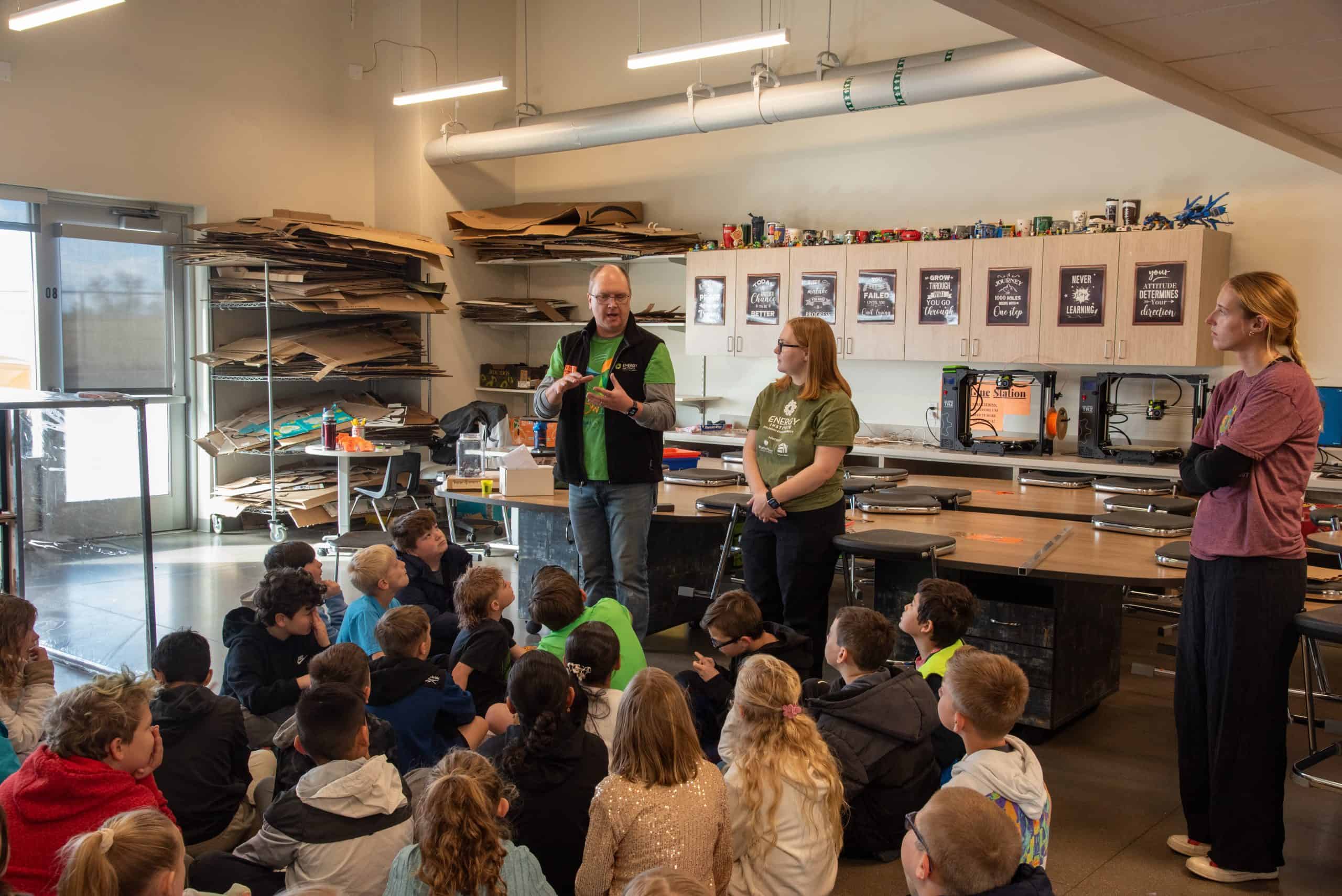 An instructor addresses a group of children sitting on the floor in a classroom filled with equipment and materials.