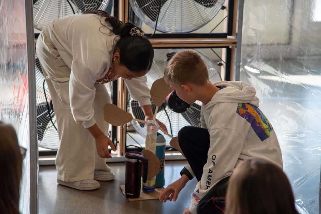 Two students are working together on a wind turbine project near large fans.