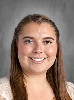 Smiling woman with long brown hair, wearing a light-colored top, against a gray background.