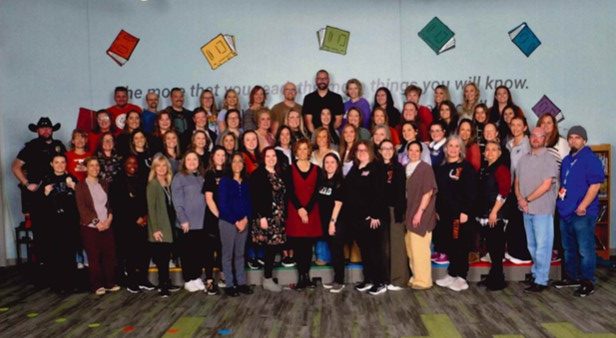 A large group of diverse adults poses together in a colorful room with books on the walls, smiling for the photo.